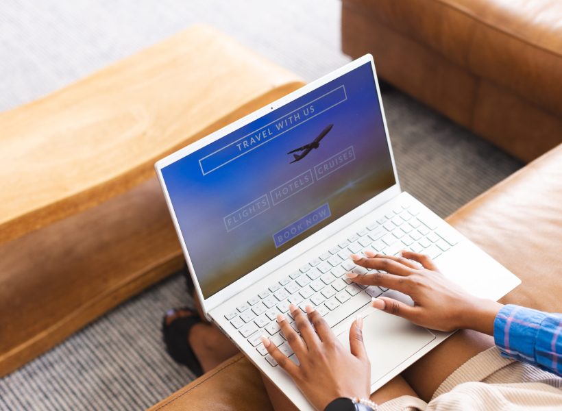 A person is booking travel online, fingers poised over a laptop keyboard. The screen displays options for flights, hotels, and cruises, suggesting vacation planning.